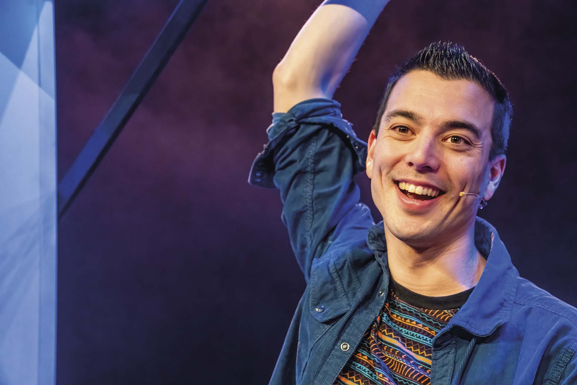 Andries Tunru staat op een podium met een headsetmicrofoon en steekt zijn arm omhoog terwijl hij lacht.