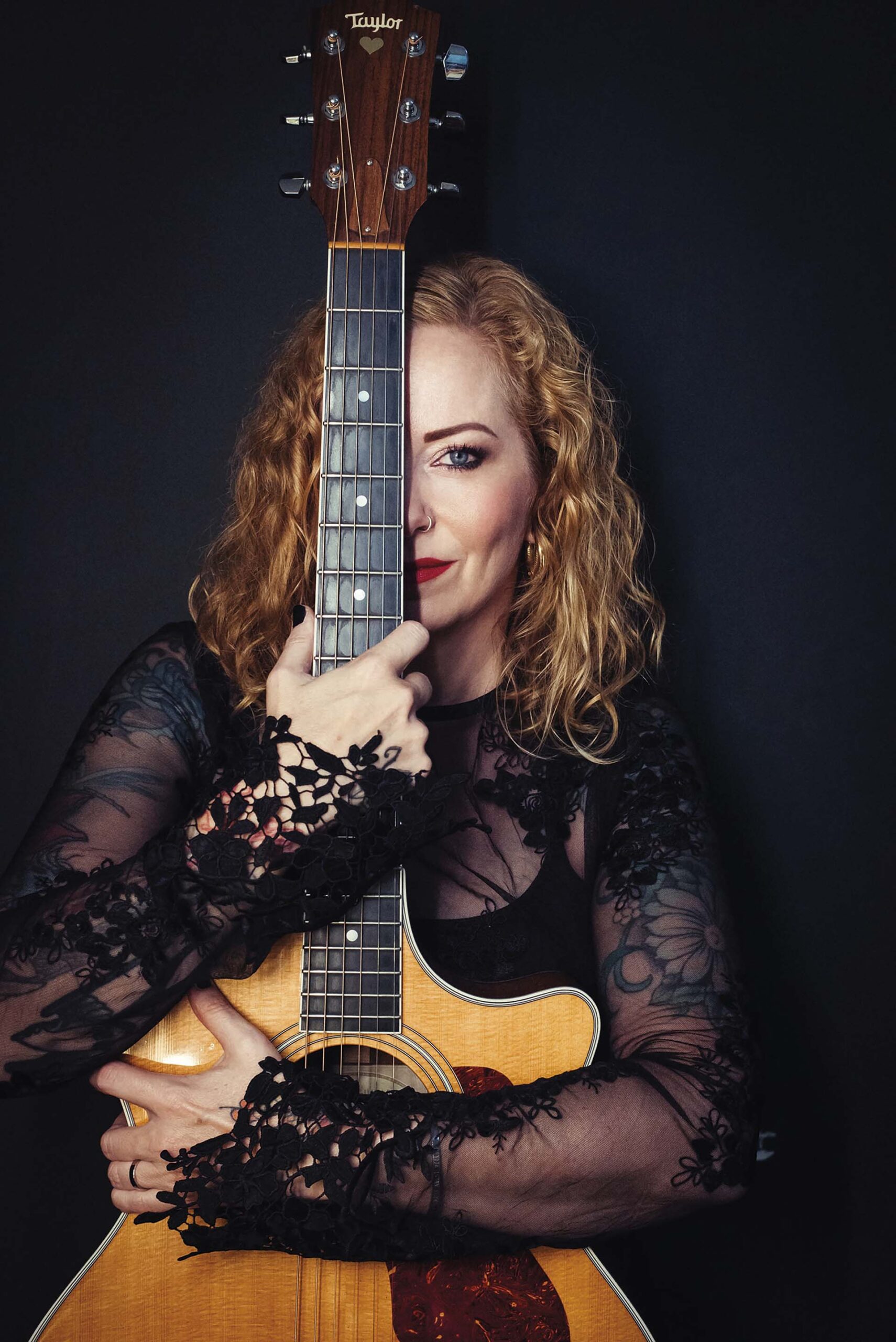 Anneke van Giersbergen houdt een akoestische gitaar voor zich, waarbij de hals haar gezicht deels bedekt, tegen een donkere achtergrond.