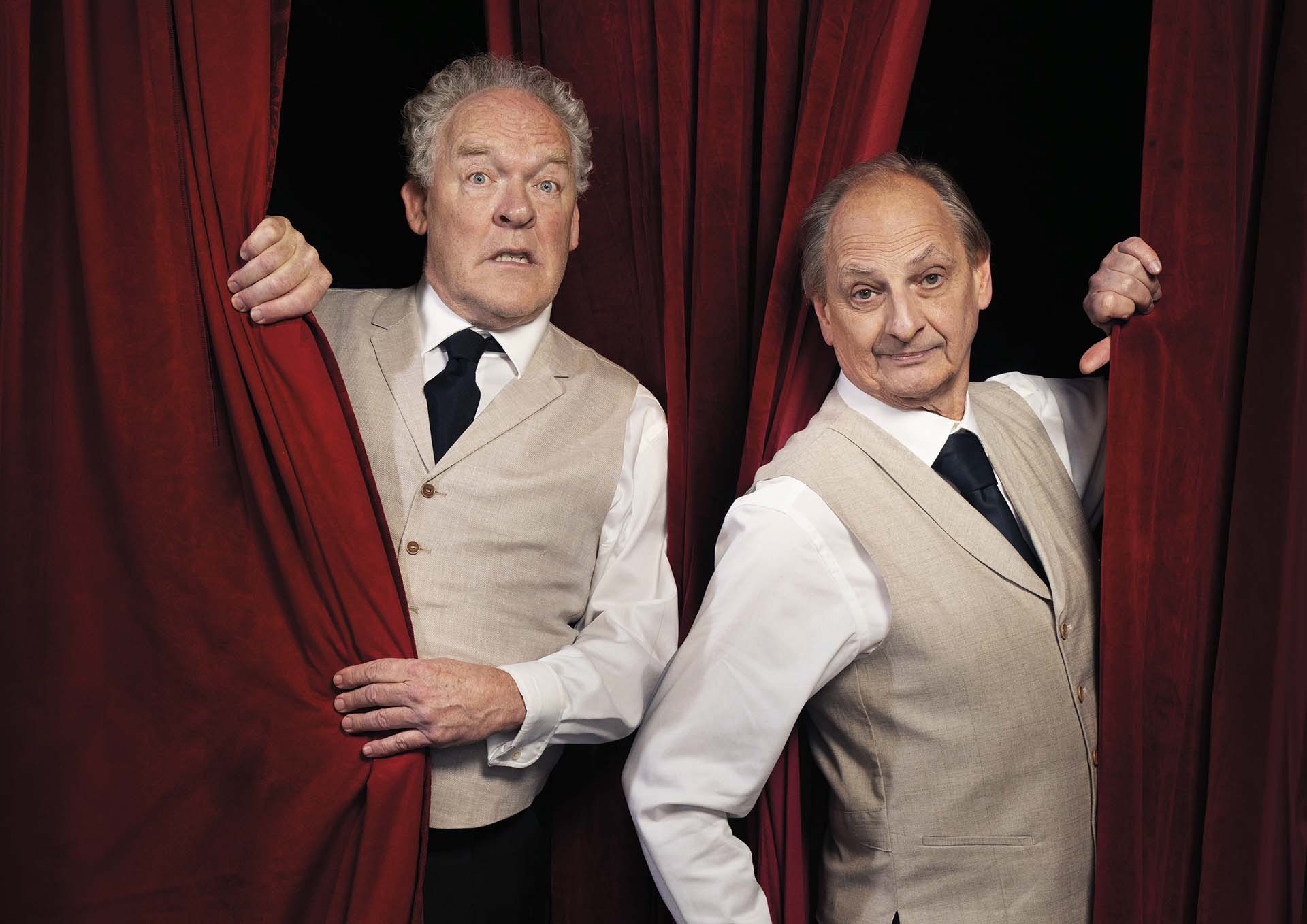 Bert Visscher en Martin van Waardenberg kijken speels door rode theatergordijnen, gekleed in witte overhemden, gilets en stropdassen, alsof ze op het punt staan het podium op te gaan
