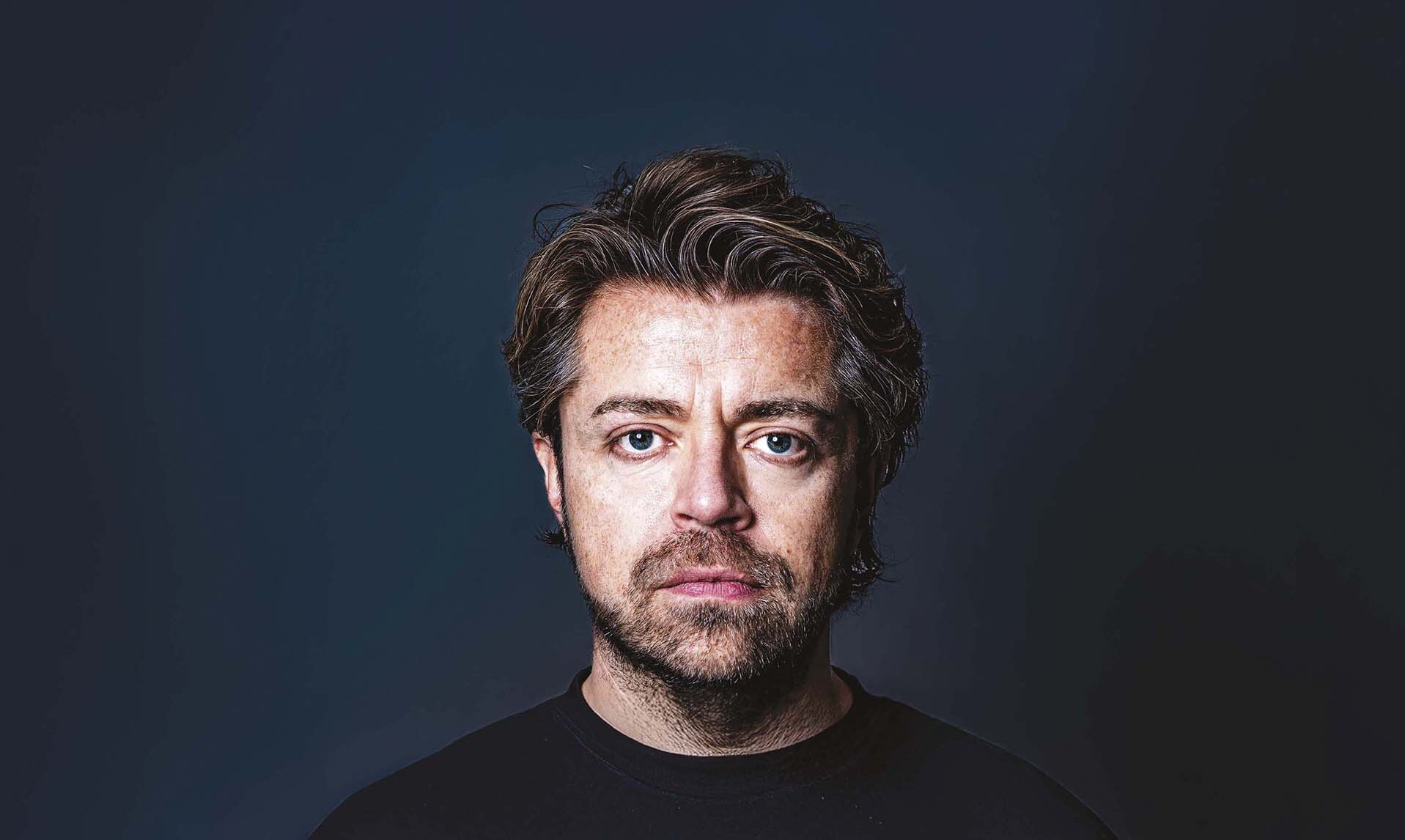 Portret een man met baard en halflang haar tegen een donkere blauwe achtergrond, recht in de camera kijkend.