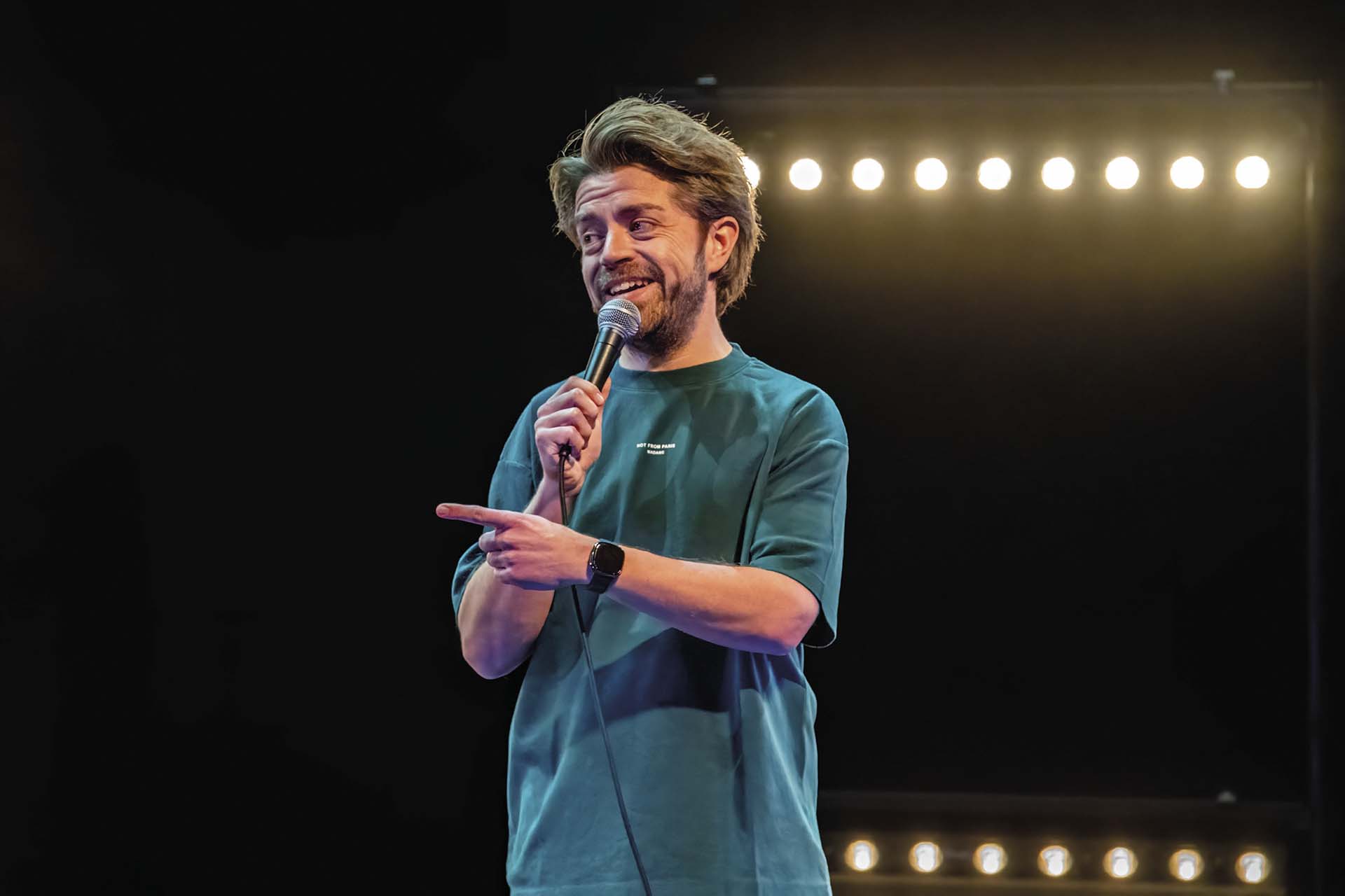 Een man staat op een podium met een microfoon in zijn hand en wijst opzij, terwijl er een rij felle podiumlampen achter hem brandt.