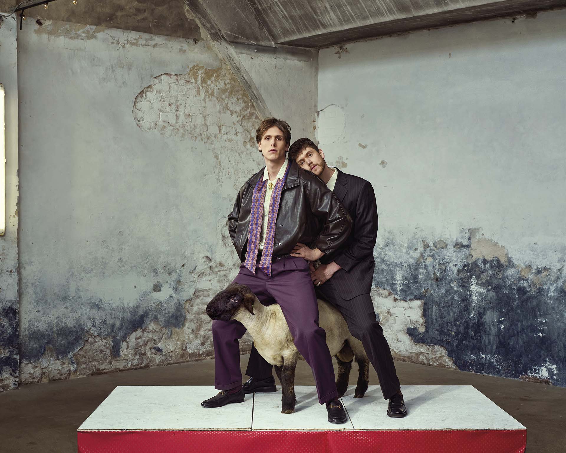 Twee mannen poseren in een lege, verweerde ruimte; één zit op een schaap terwijl de ander achter hem staat en zijn hoofd tegen hem aan leunt.