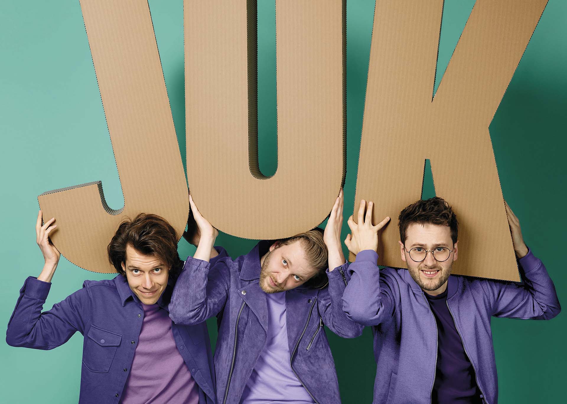 Drie mannen in paarse kleding houden grote kartonnen letters vast boven hun hoofd die samen JUK vormen, tegen een groenblauwe achtergrond.