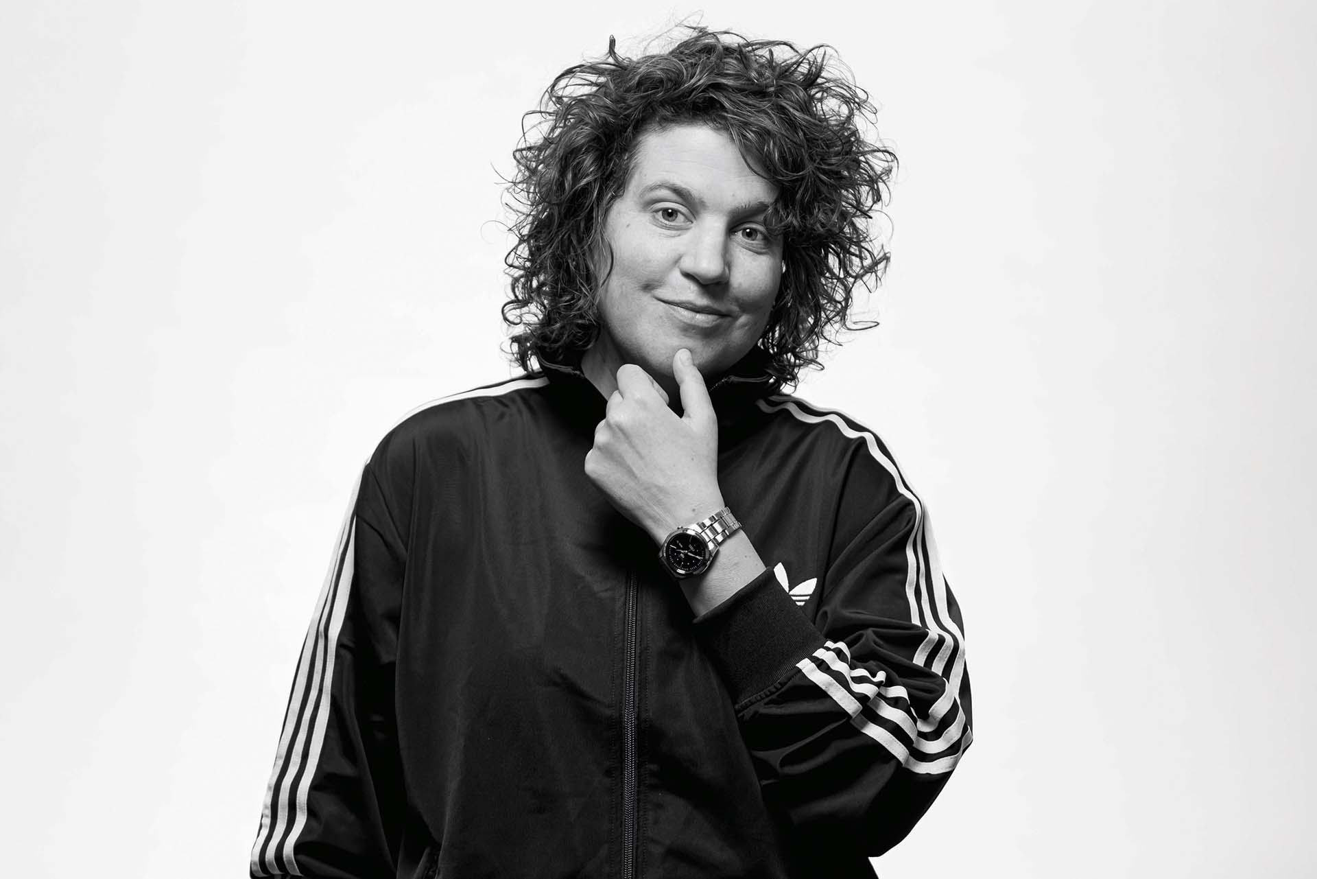 Kim Schuddeboom in zwart-wit portret met krullend haar en sportief jasje, glimlacht licht met hand onder haar kin