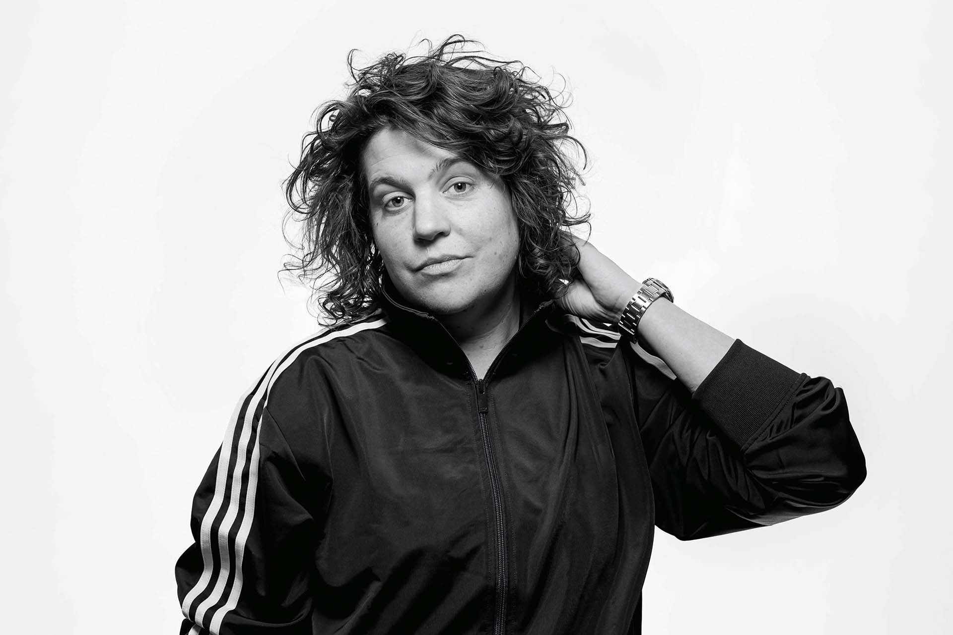 Kim Schuddeboom in zwart-wit portret met krullend haar en sportief jasje, kijkt recht in de camera met hand in haar nek