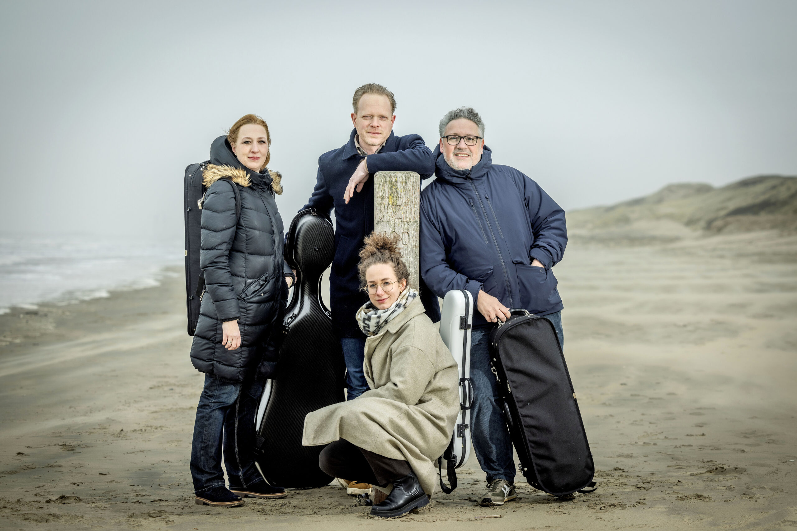 Matangi Quartet op het strand met instrumentkoffers, ontspannen pose in een open kustlandschap