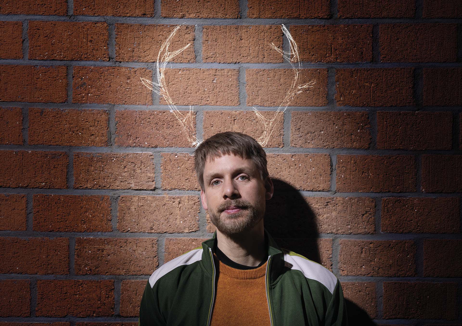 Een man staat voor een bakstenen muur en kijkt in de camera, met achter zijn hoofd getekende geweien op de muur.