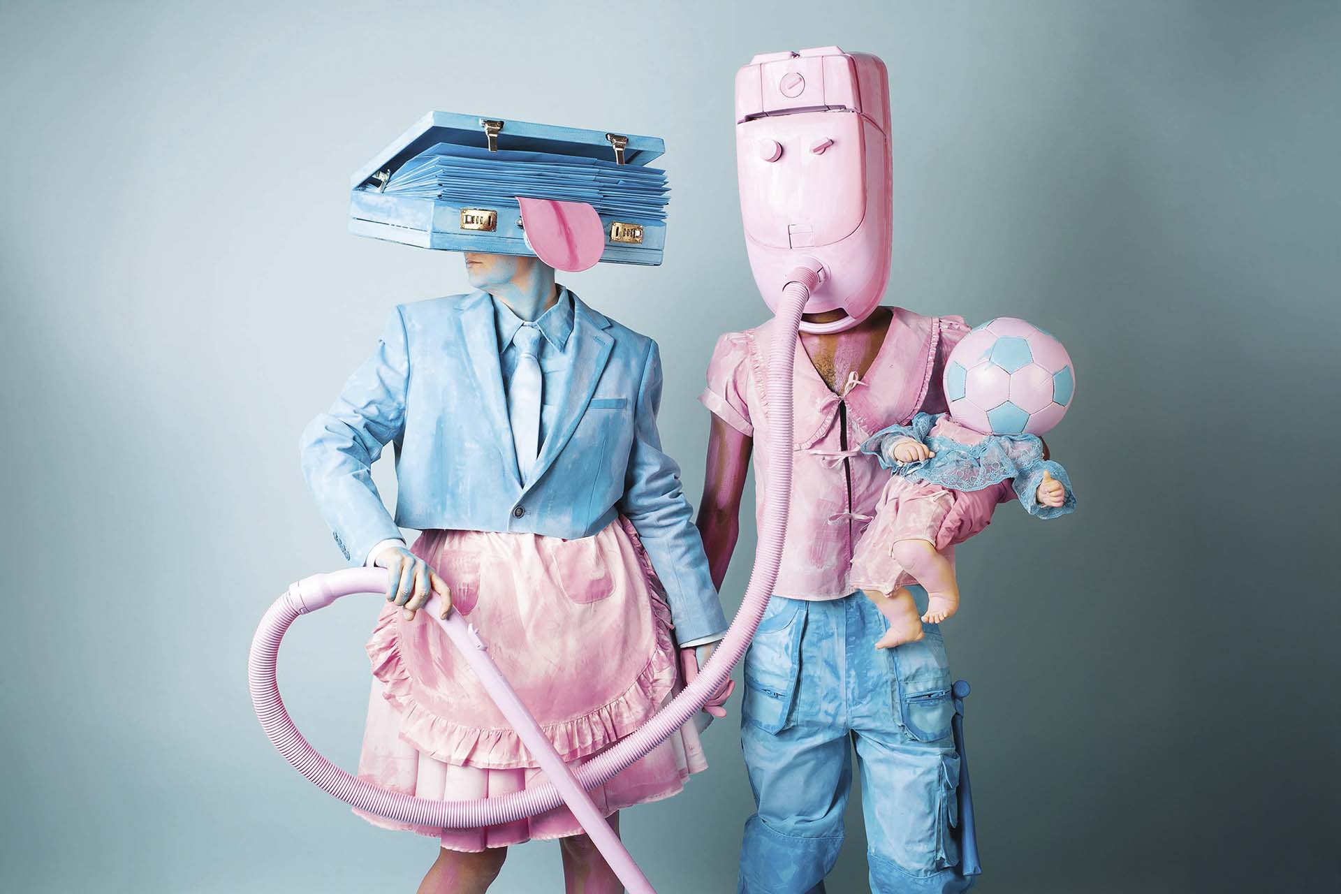 Twee performers in pastelkleuren dragen surrealistische kostuums met objecten als hoofd en poseren met een stofzuigerslang en een pop tegen een zachte achtergrond