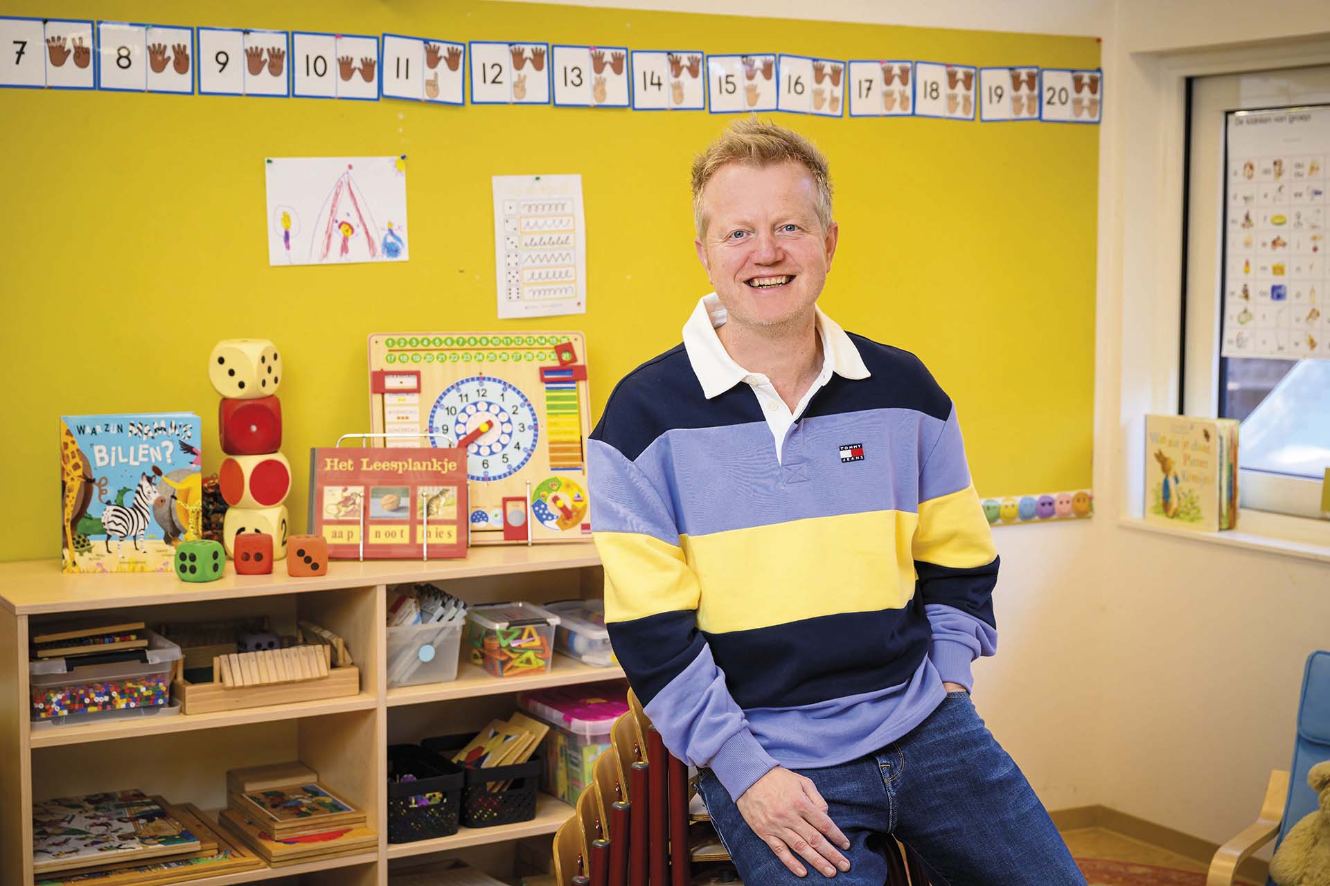 Meester Mark zit lachend in een kleurrijk klaslokaal, omringd door educatief speelgoed, boeken en telmateriaal tegen een gele muur