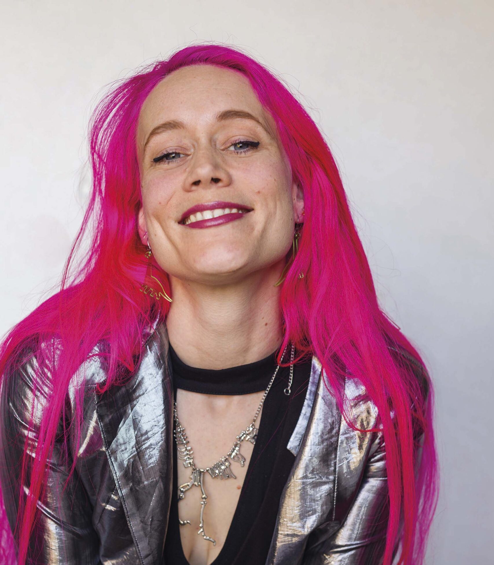 Melanie met felroze haar en zilveren jasje kijkt glimlachend in de camera tegen een lichte achtergrond