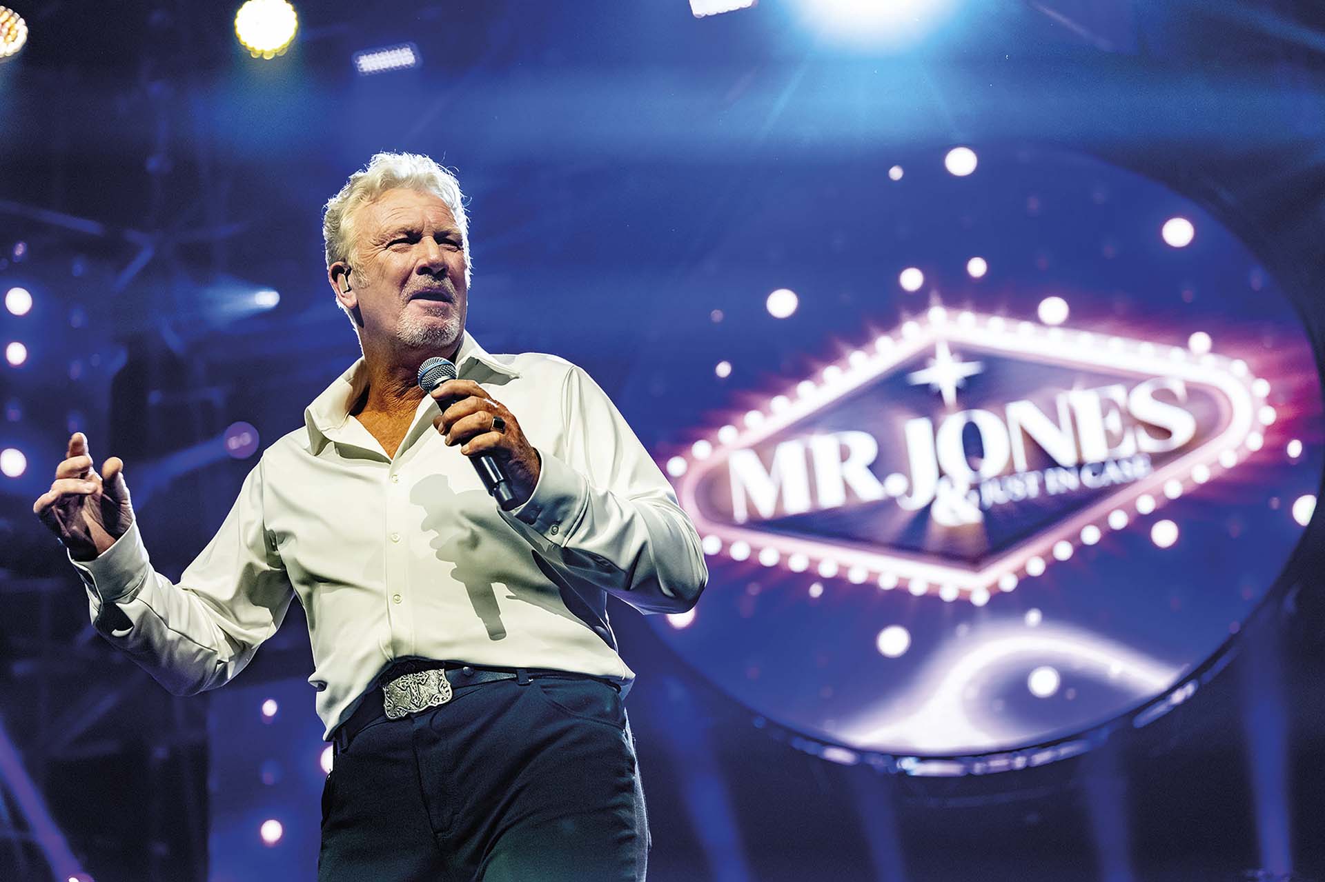 Mr. Jones zingt op podium met microfoon, omringd door podiumlichten en groot verlicht logo op achtergrond in concertsetting