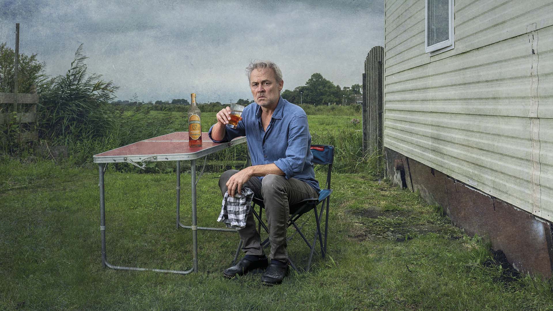 Marcel Hensema zit naast een caravan aan een campingtafel met een glas drank, in een landelijke omgeving onder een bewolkte lucht.