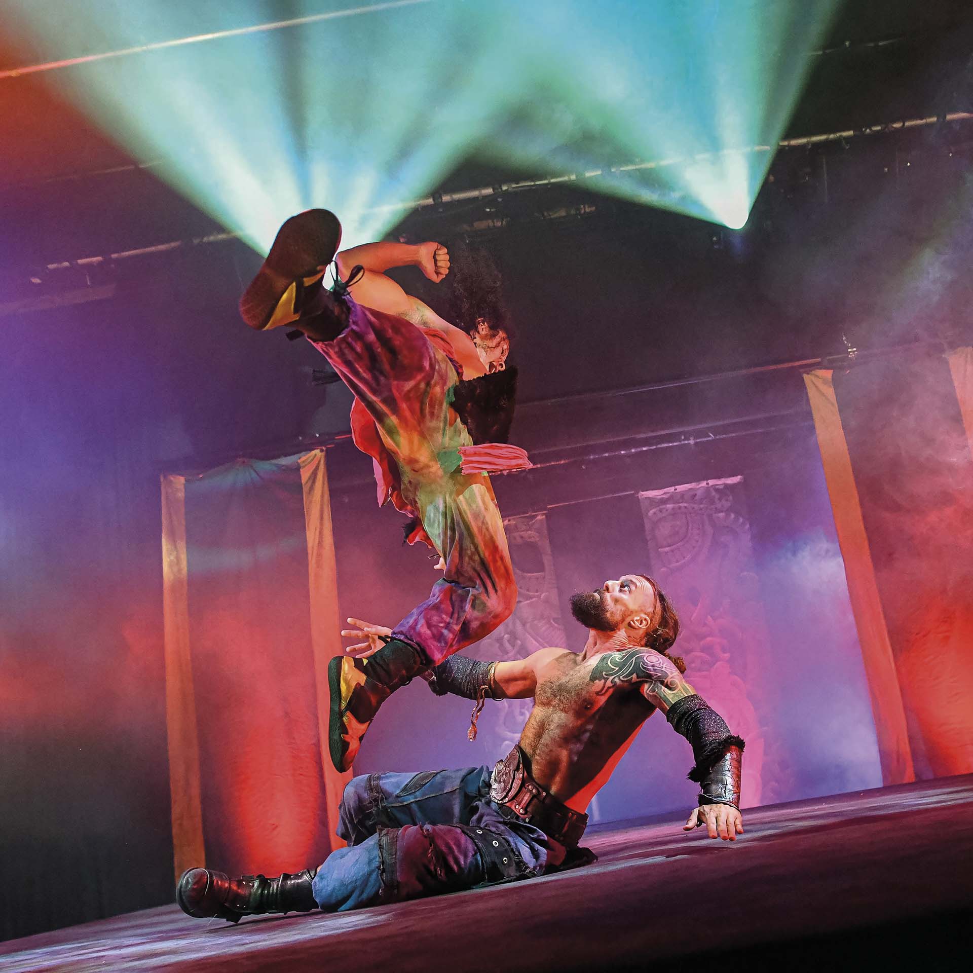 Acrobatische scène waarbij een performer een hoge trapbeweging maakt boven een liggende man op een verlichte theatervloer