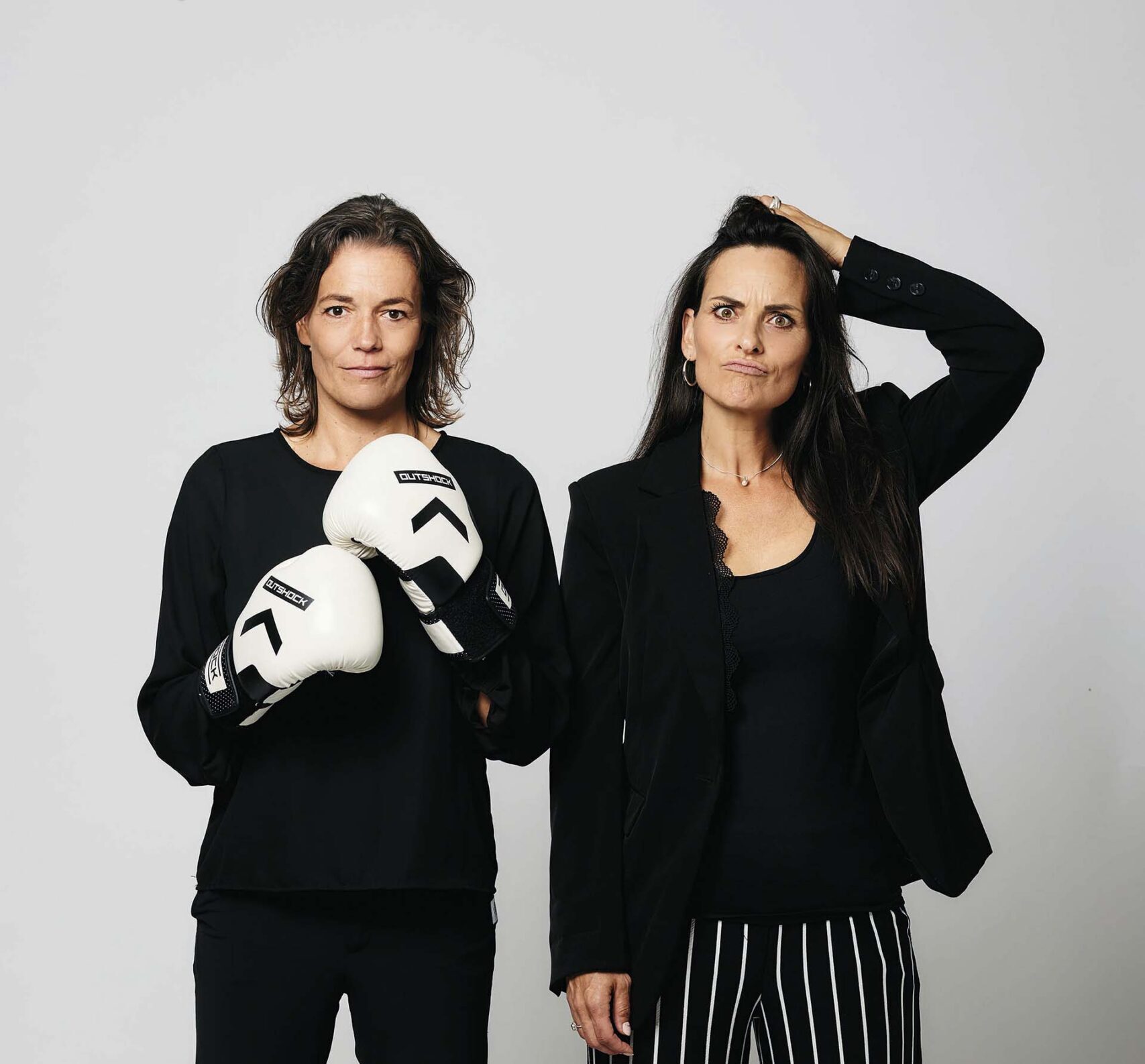 Ilse van den Heuvel en Marieke van Ginneken staan naast elkaar tegen een neutrale achtergrond; Ilse draagt bokshandschoenen, terwijl Marieke met een vragende blik haar hand in haar haar legt