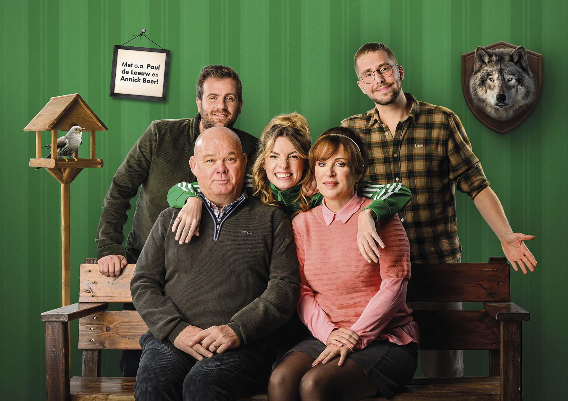 Paul de Leeuw, Annick Boer e.a. poseren samen op een bank tegen een groene achtergrond met decorstukken zoals een vogelhuisje en een opgezette wolf