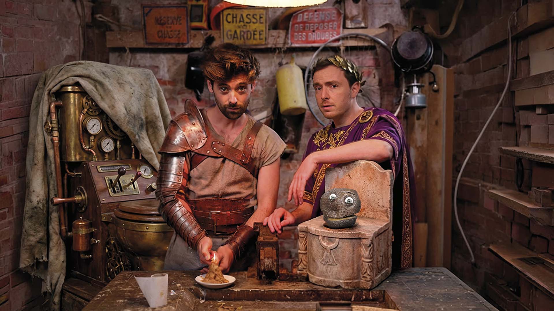 Pieter en Willem in kostuumachtige outfits staan in een rommelige werkplaats met oude machines en rekwisieten