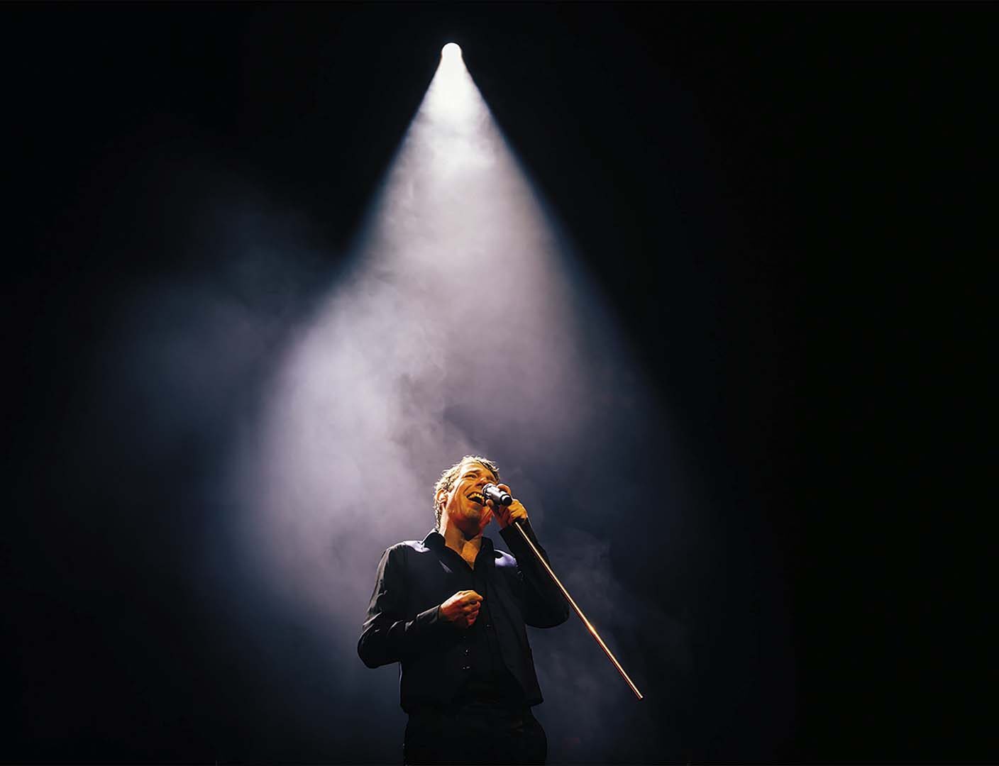 Zanger staat alleen onder een felle spot op donker podium en zingt in microfoon, omringd door rook en theatrale belichting