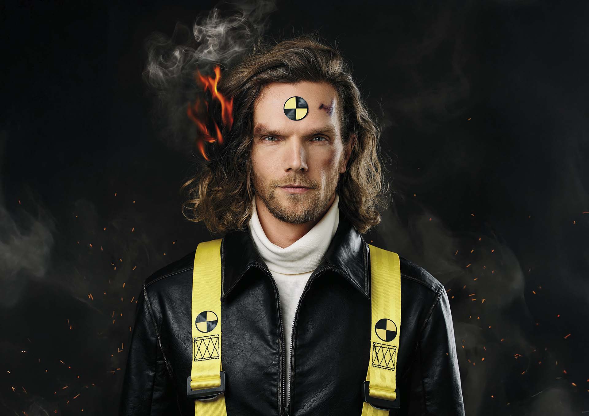 Portret van Ruud Smulders met lange haren en leren jas, met brandwerende banden en testmarkering op zijn voorhoofd, tegen donkere achtergrond met rook en vuursporen