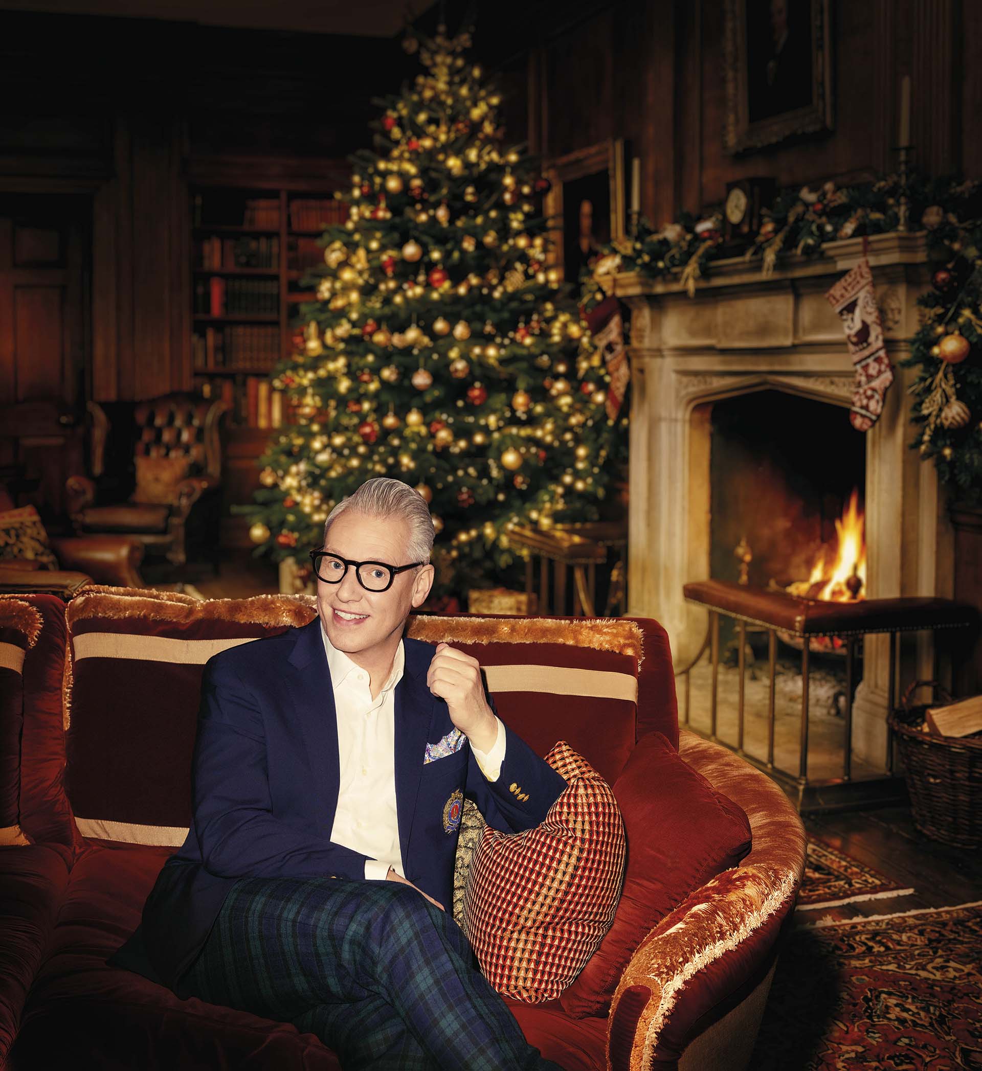 Rudolph van Veen zit op een bank in een sfeervolle woonkamer met een versierde kerstboom en brandende open haard op de achtergrond.