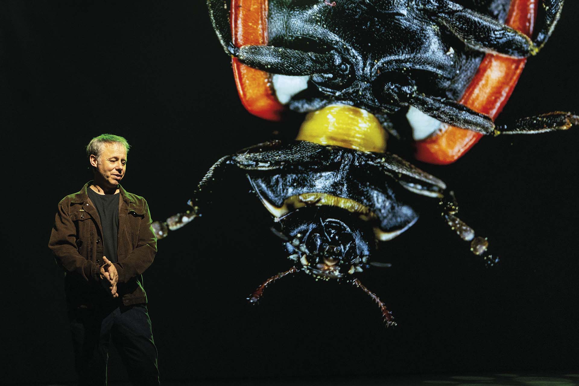 Luc Hoogenstein op podium met groot scherm waarop close-up van insect te zien is, tijdens natuurtheatercollege met donkere achtergrond