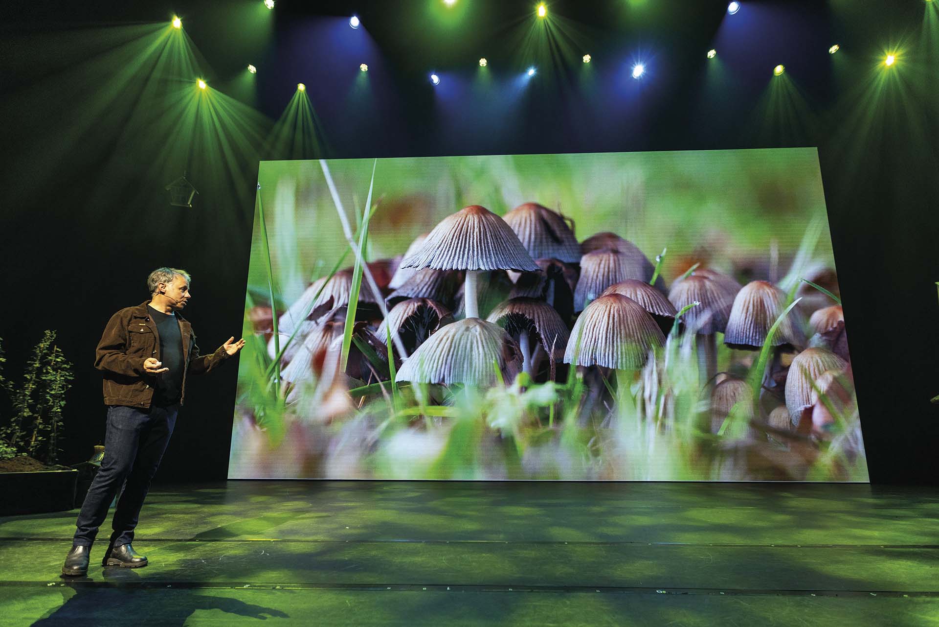 Luc Hoogenstein wijst naar groot scherm met paddenstoelenbeeld, tijdens theatercollege met groene belichting en natuurthema