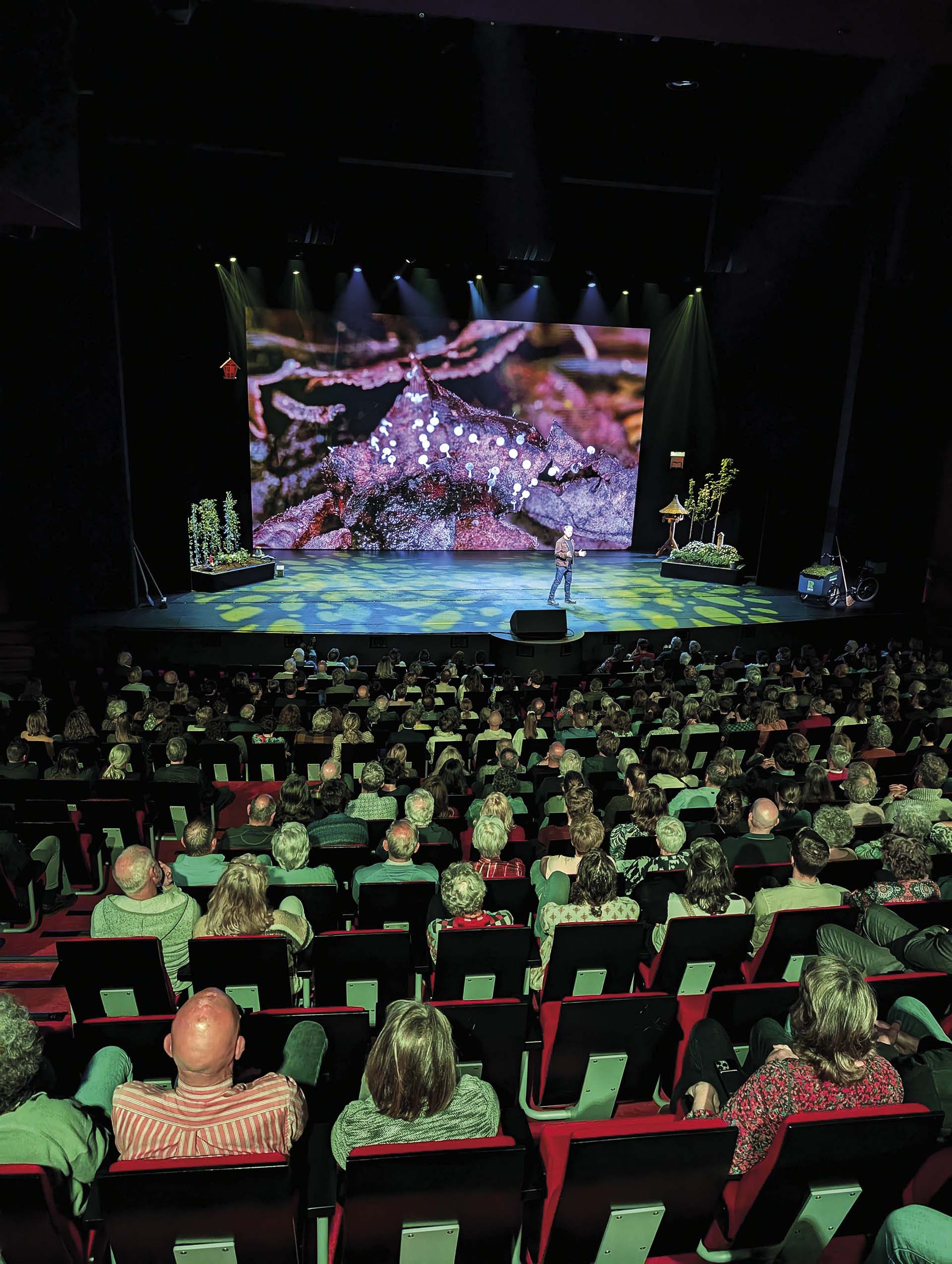 Publiek in theaterzaal kijkt naar Luc Hoogenstein op podium met groot scherm vol natuurbeelden, tijdens voorstelling Stadssafari