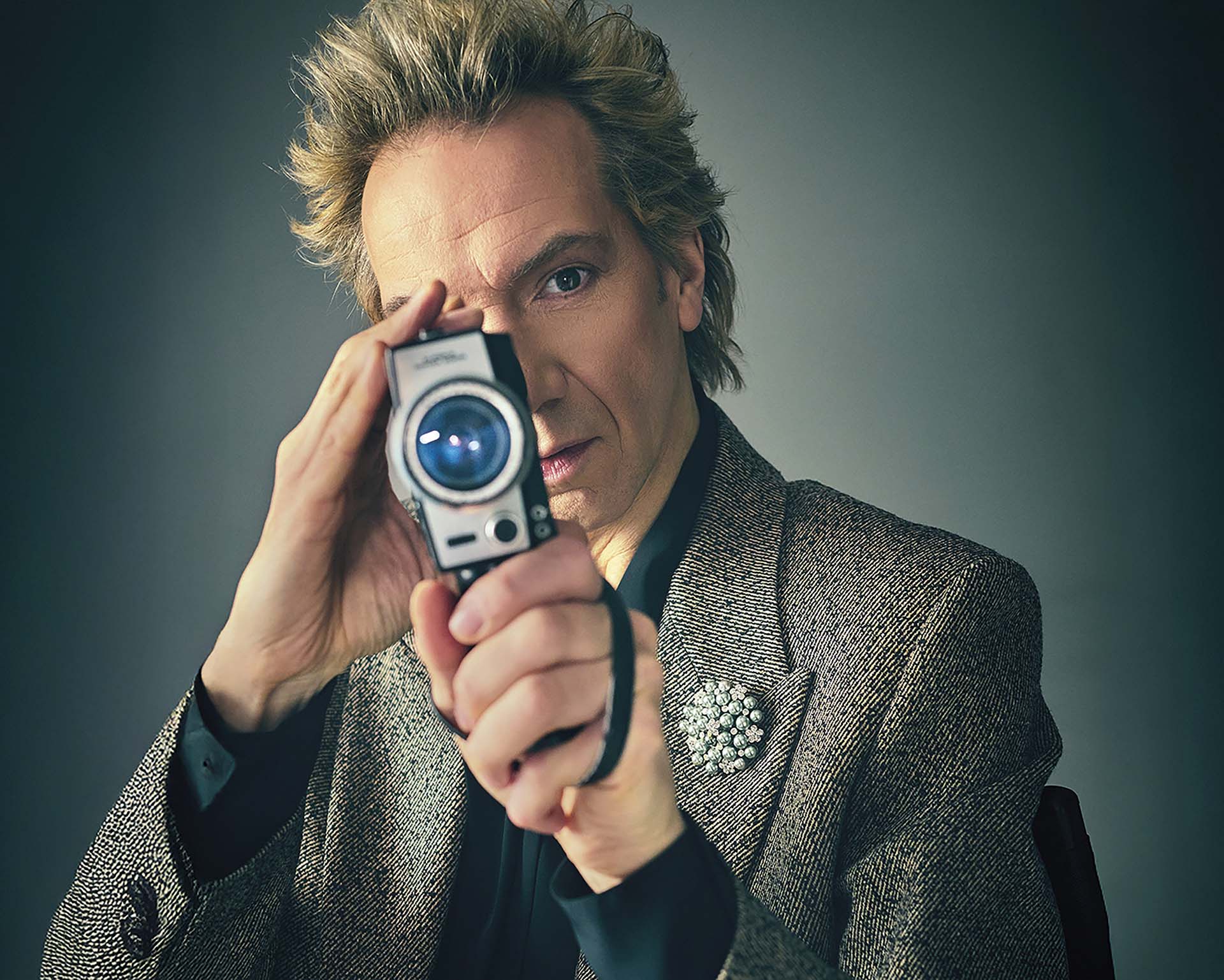 Sven Ratzke houdt een kleine camera voor zijn oog en kijkt recht in de lens, gekleed in een stijlvol jasje tegen een rustige achtergrond