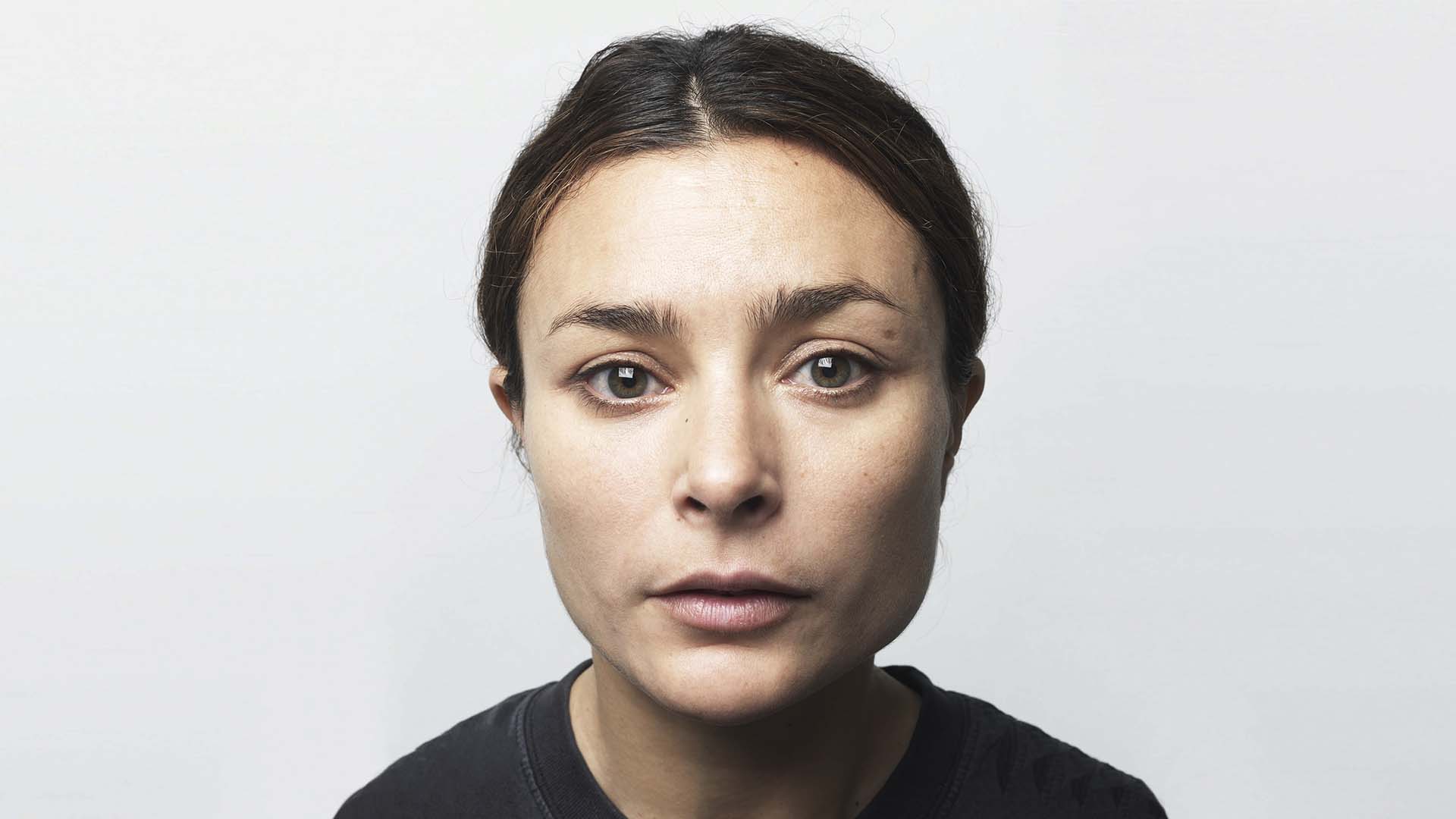 Vrouw kijkt recht in de camera met een neutrale, indringende blik tegen een lichte achtergrond