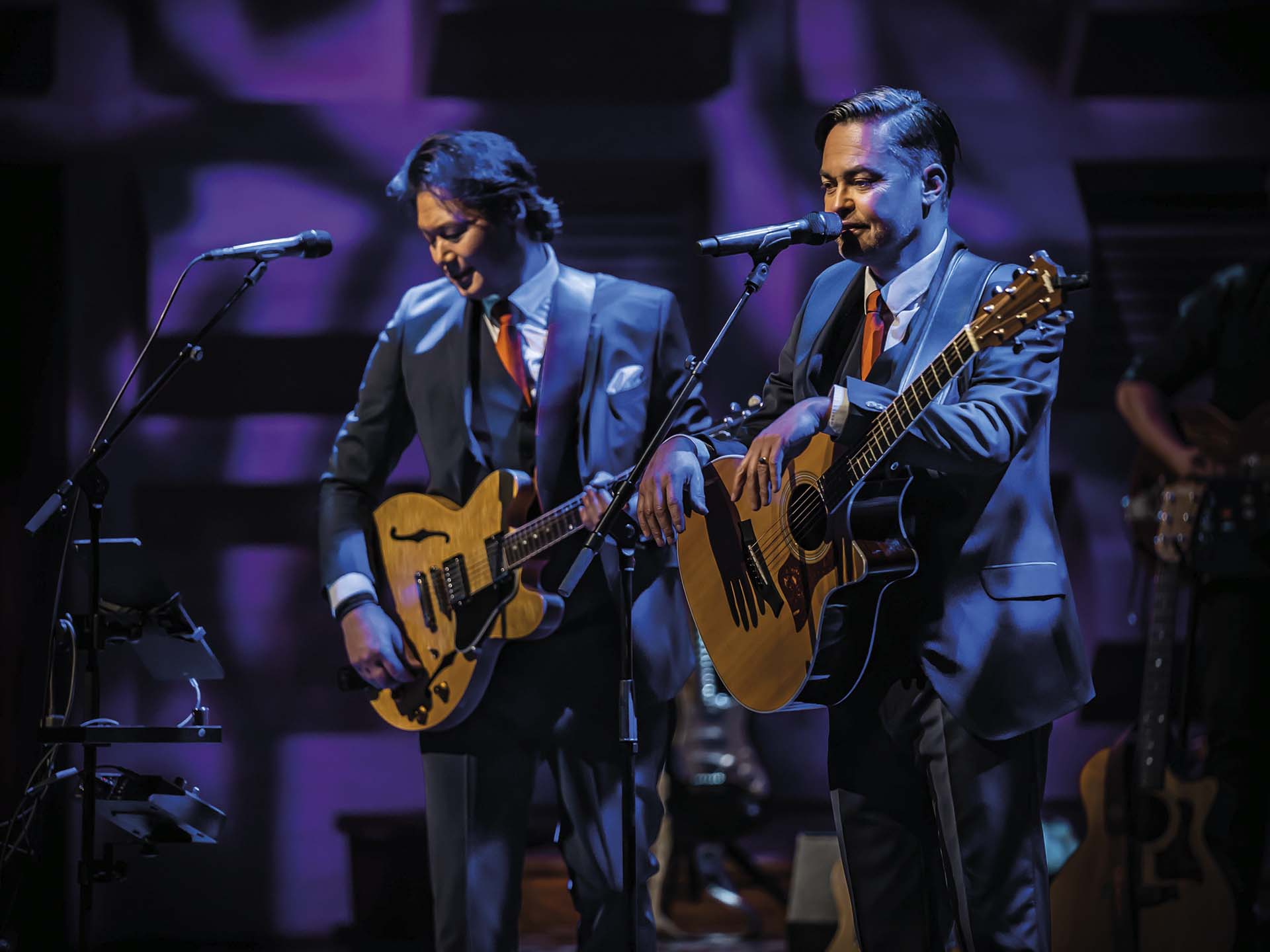 Twee muzikanten in pak spelen gitaar en zingen naast elkaar op een sfeervol verlicht podium