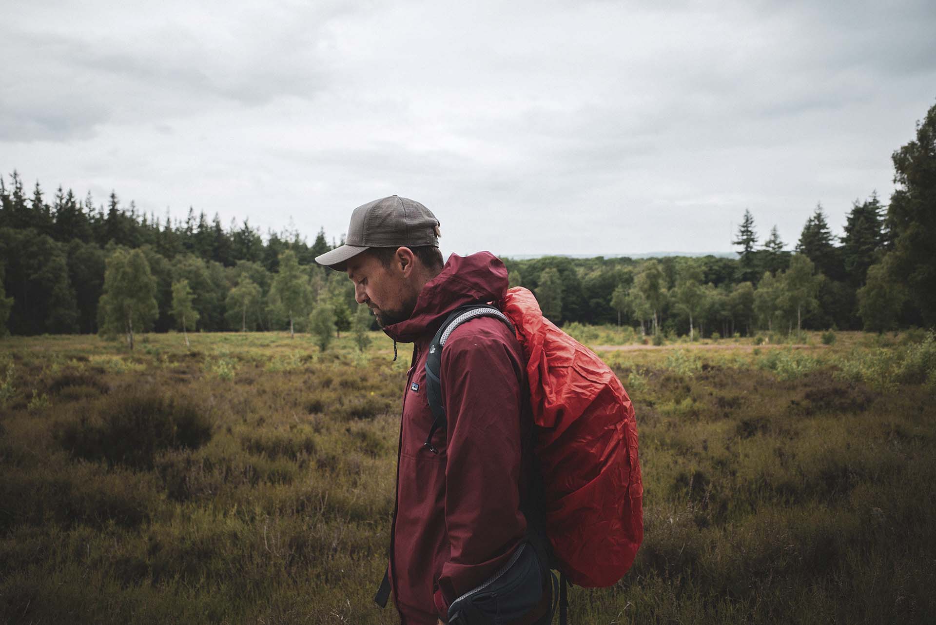Tim Knol met pet en rugzak loopt door een open natuurgebied met gras en bomen onder een bewolkte lucht
