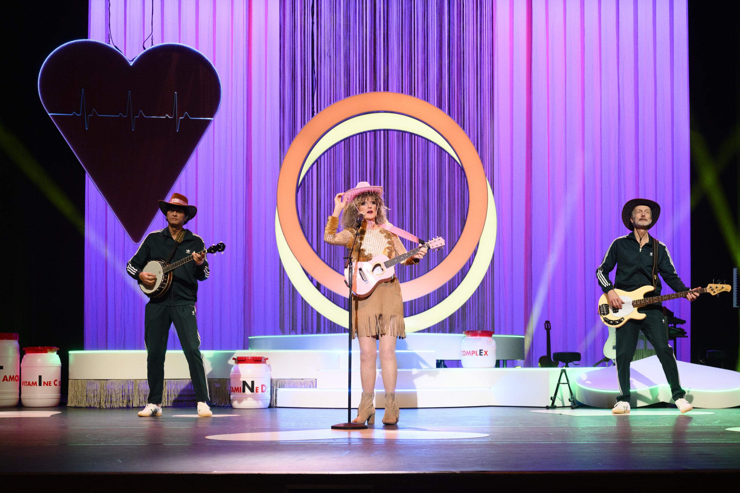 Tineke Schouten zingt met gitaar midden op het podium, geflankeerd door twee muzikanten, tegen decor met hartvorm en ronde lichtinstallatie