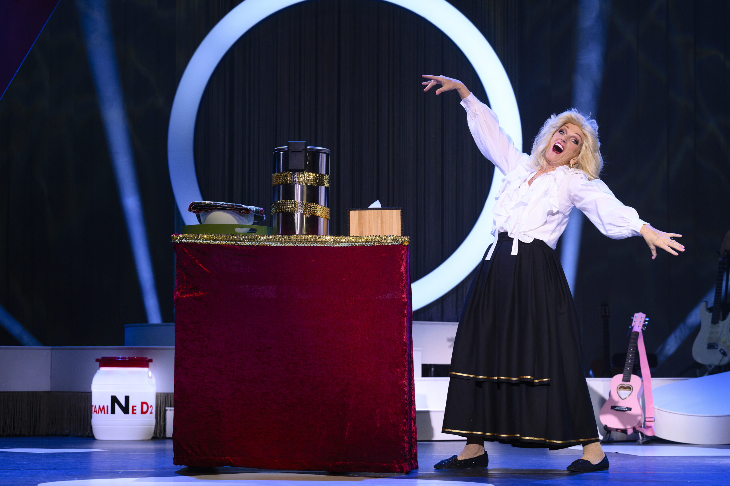 Tineke Schouten op het podium naast een tafel met attributen, in zwarte rok en witte blouse, met expressieve pose tegen decor met ronde lichtvorm