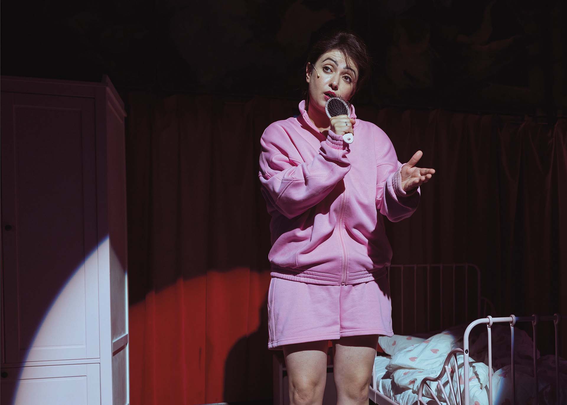Vrouw in roze outfit staat op toneel en zingt in haarborstel als microfoon, naast kinderbed en kast in theatrale setting met rood licht