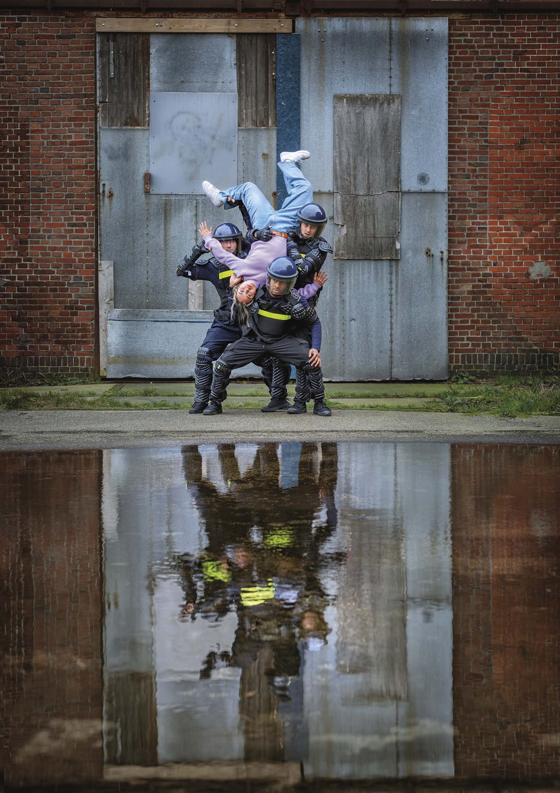 Drie agenten in oproeruitrusting houden een persoon ondersteboven vast voor een industriële muur, met hun reflectie zichtbaar in het water ervoor