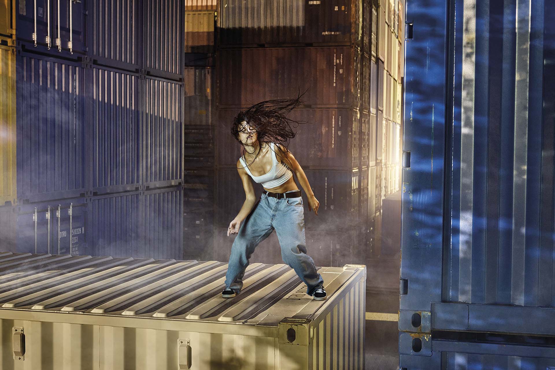 Danser beweegt krachtig op een zeecontainer tussen hoge stapels containers, met wapperend haar en industriële setting
