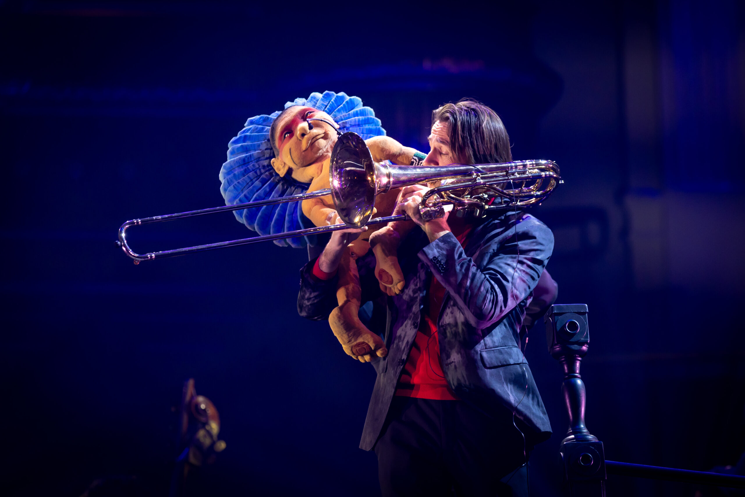 Muzikant bespeelt een trombone op het podium terwijl een grote pop met blauwe kraag over zijn schouder meekijkt