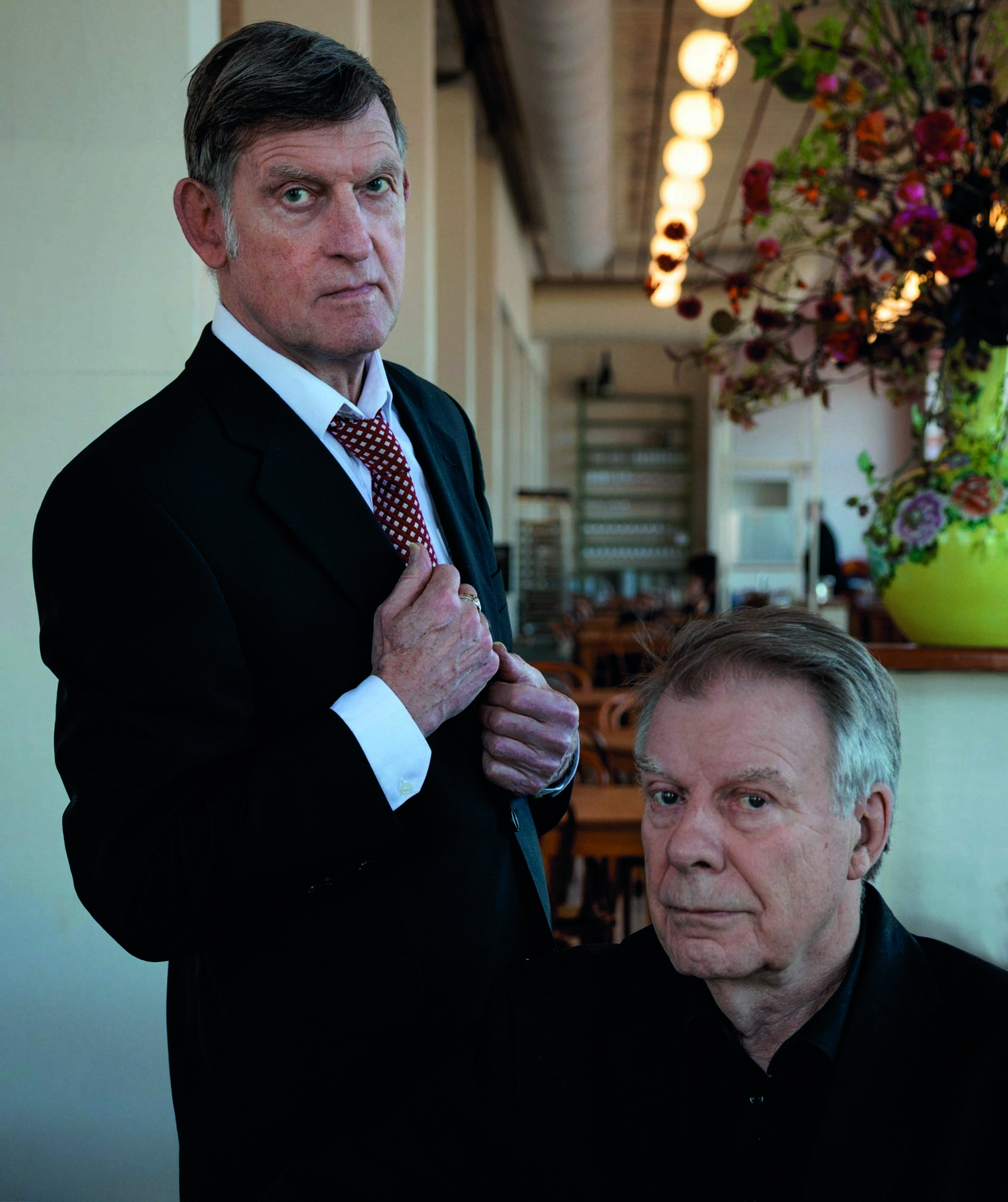 Huub van der Lubbe en Jan Robijns poseren in een sfeervol café-interieur met bloemen en hanglampen op de achtergrond