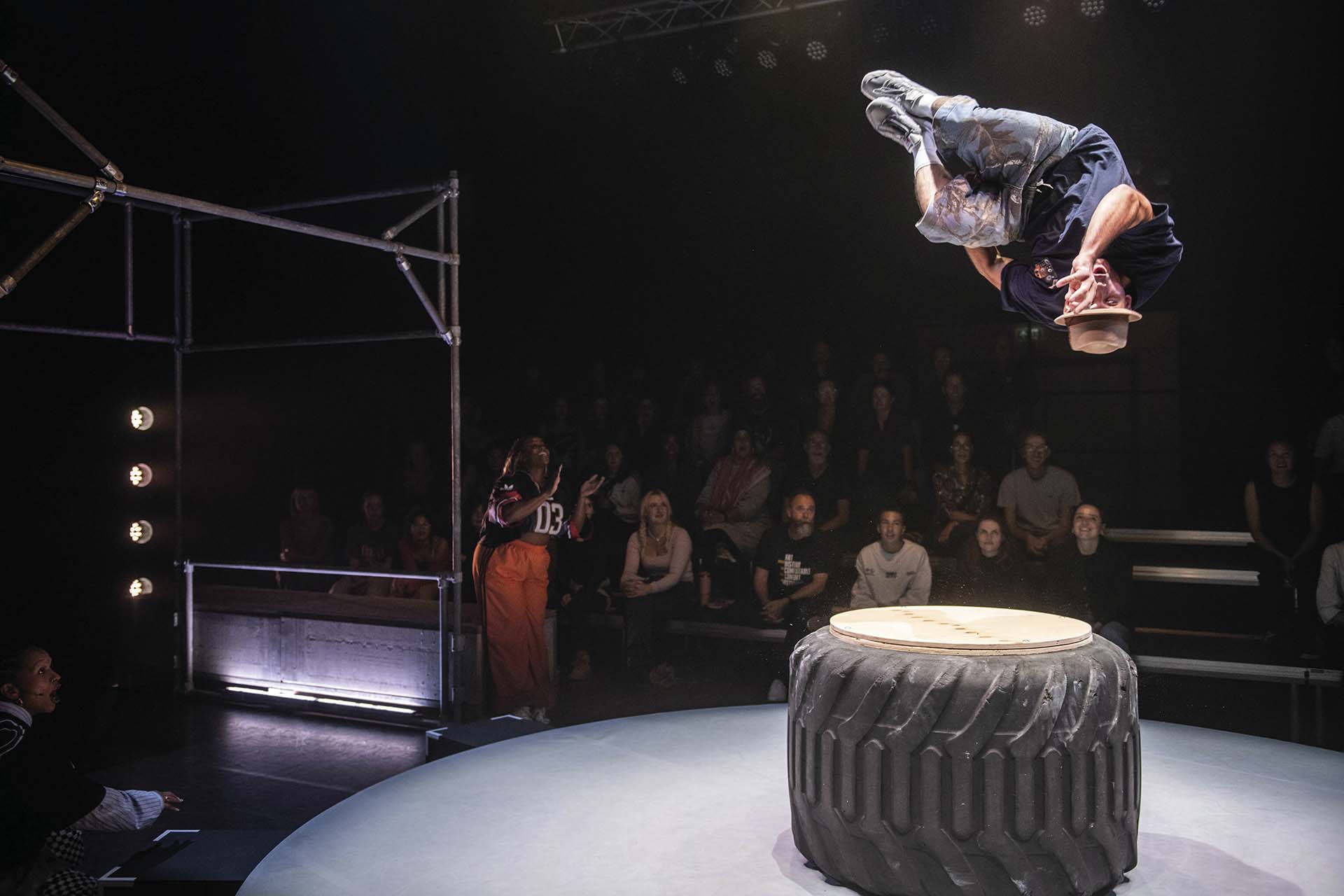 Danser maakt salto boven ronde speeltafel, publiek kijkt toe
