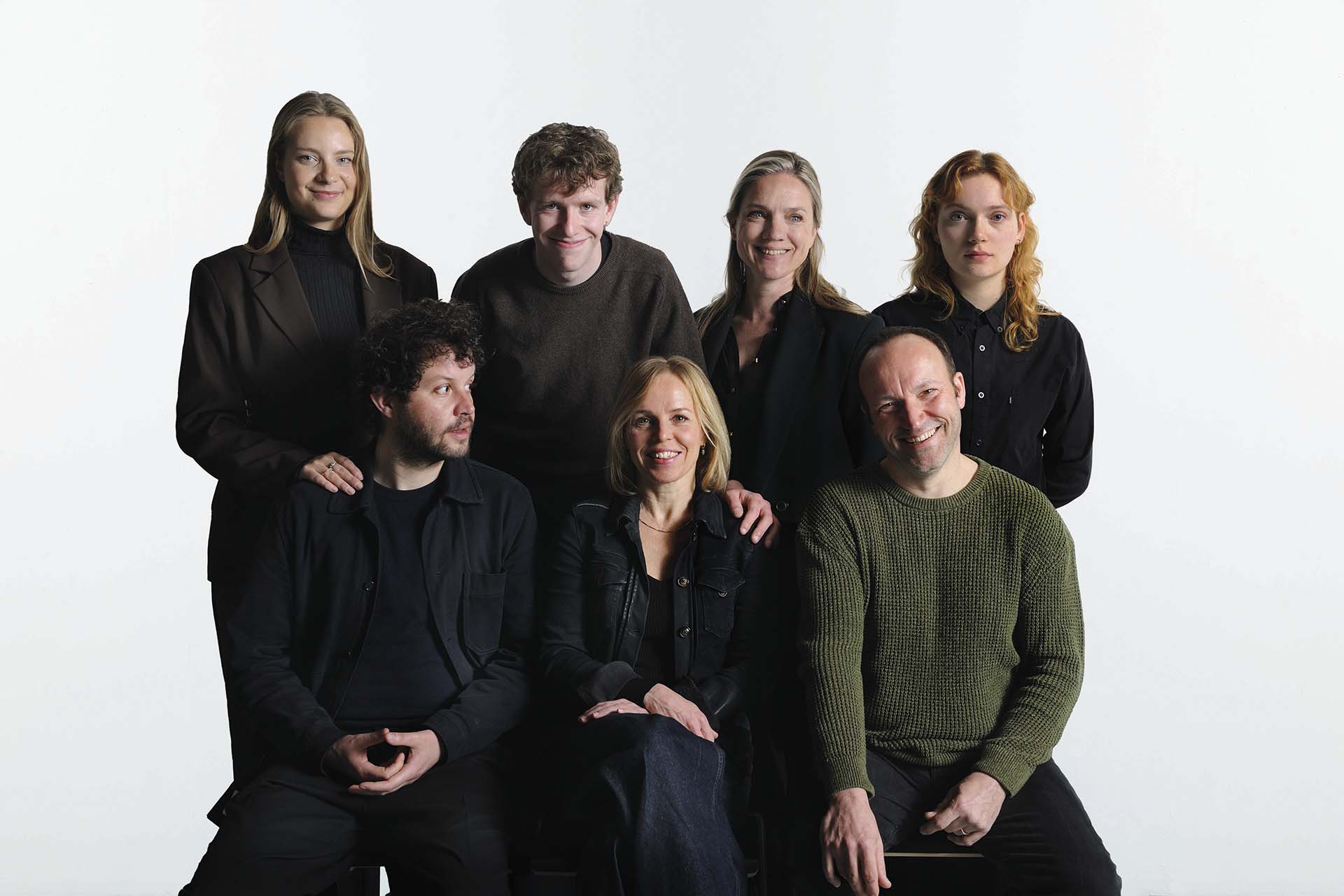Een groep van zeven mensen poseert samen voor een neutrale achtergrond. Drie zitten vooraan en vier staan erachter. Ze zijn casual en donker gekleed en kijken ontspannen en vriendelijk in de camera, alsof het een cast- of teamfoto is.