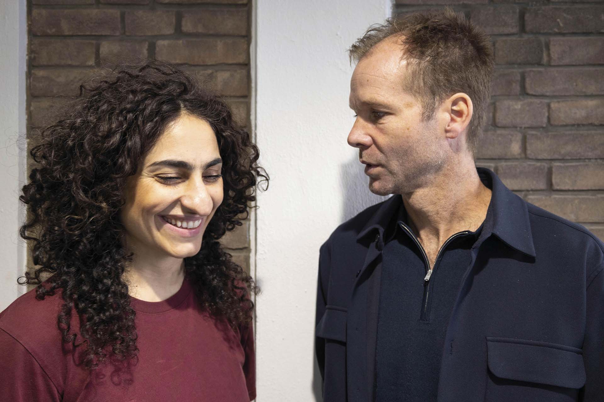 Twee spelers van Wunderbaum staan naast elkaar voor een bakstenen muur, terwijl de man de glimlachende vrouw aankijkt