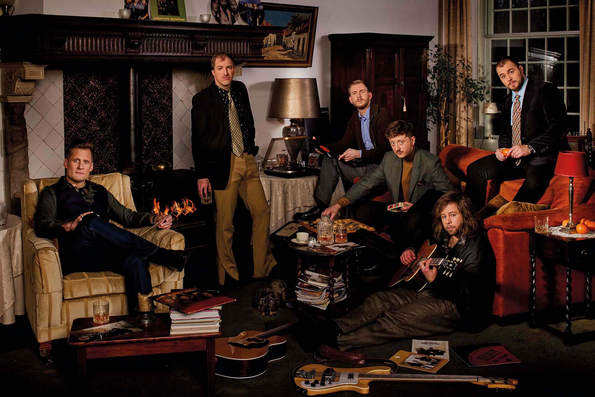 Zes mannen in stijlvolle woonkamer met open haard, instrumenten en drankjes, poseren ontspannen in vintage setting met gitaar, platen en warme verlichting