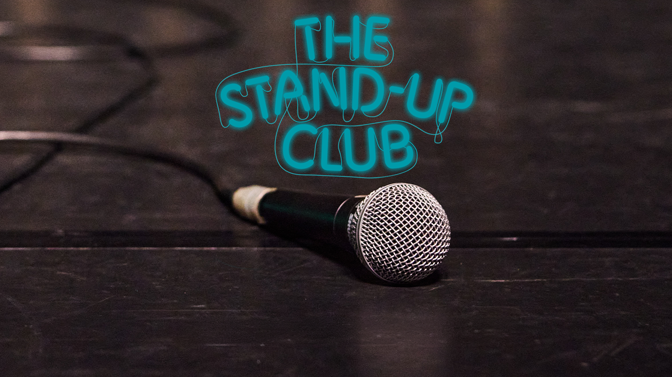 Een microfoon op een donker podium met op de achtergrond de tekst ‘The Stand-Up Club’ in blauwe neonletters.